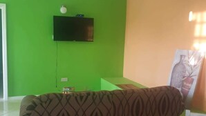 A living room with a green wall, a flat-screen TV mounted above a glass coffee table, and a patterned sofa. - Corinth Convinced Apartment One (Gros Islet)