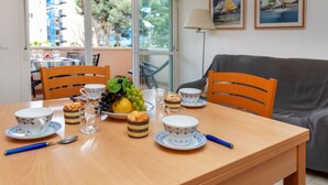 A dining table set with plates, cups, and a fruit bowl, with a view of the outside through large windows. - Cosy apartment for 4 people with WIFI, pool, TV and terrace (Salou)
