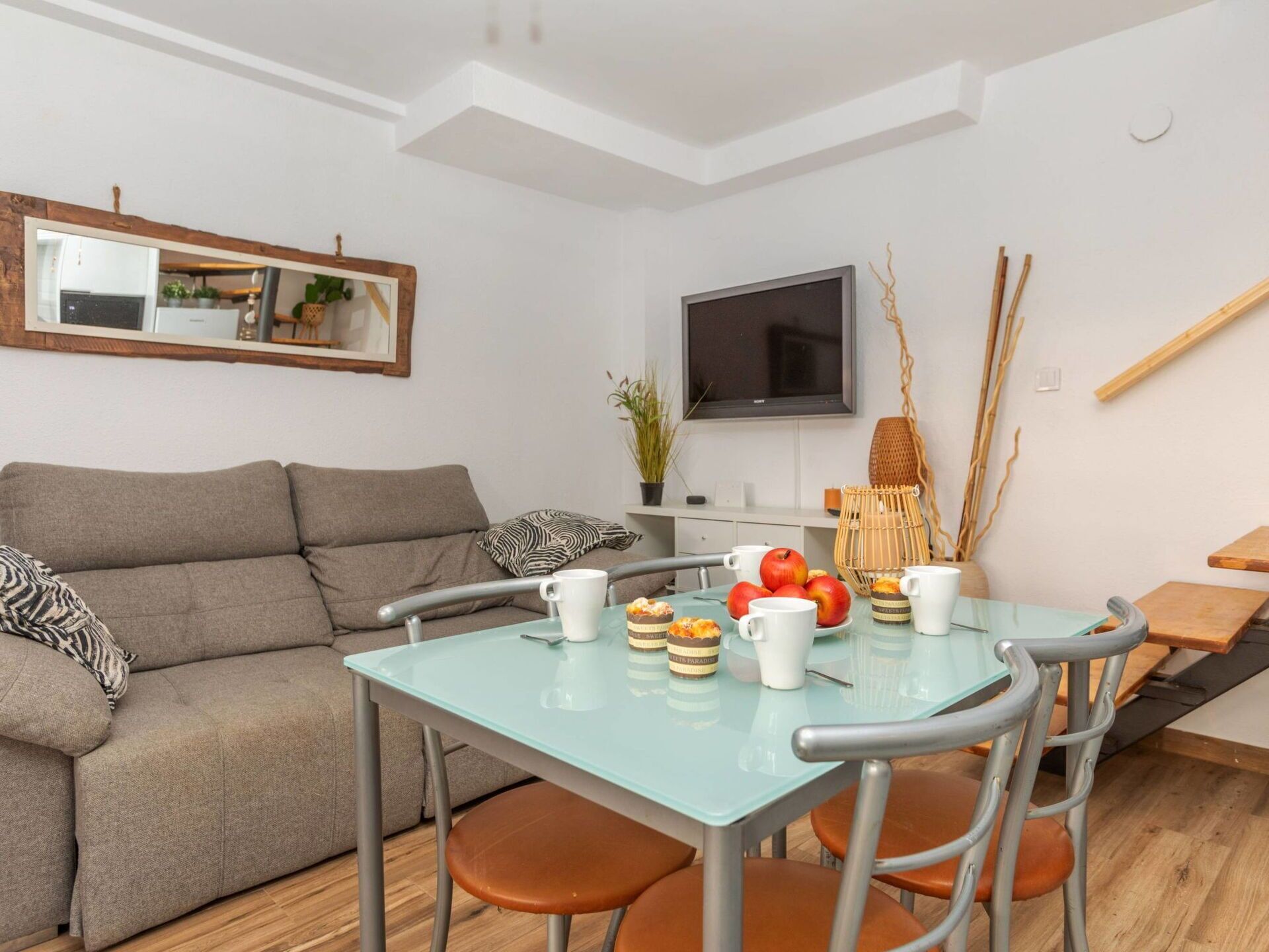 A modern living room with a glass dining table, grey sofa, and a flat-screen TV mounted on the wall.