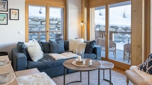 A cozy living room with a blue sofa, a coffee table, and a view of a snowy outdoor area through large windows. - Cosy apartment for 6 guests with hot tub, WIFI, TV and balcony (Basse-nendaz)