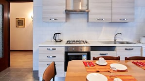 Una cocina moderna con armarios de madera clara, una estufa de acero inoxidable y un lavabo. Hay una mesa de comedor preparada con platos, cubiertos y una servilleta.