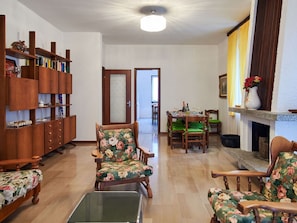 A living room with a wooden bookshelf, floral armchairs, a glass coffee table, and a dining area with a table set for four. - Beautiful apartment in villa for 16 people with WIFI, TV, balcony and pets allowed (Dervio)