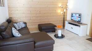 A modern living room with a brown leather sofa, a glass coffee table with oranges, a TV on a wooden cabinet, and a floor lamp. - Beautiful apartment for 4 guests with WIFI, TV, terrace and panoramic view (Evolène)