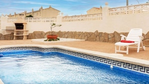 A swimming pool with a tiled edge, a barbecue area, and a white chair with a red towel. - Beautiful private villa for 6 guests with private pool, TV and patio (Miami Platja)
