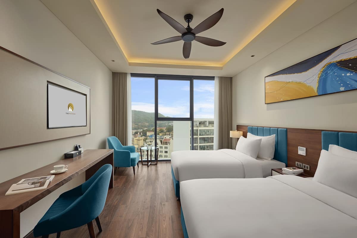 A hotel room with a desk, two beds, a ceiling fan, a large window with a city view, and a painting on the wall.