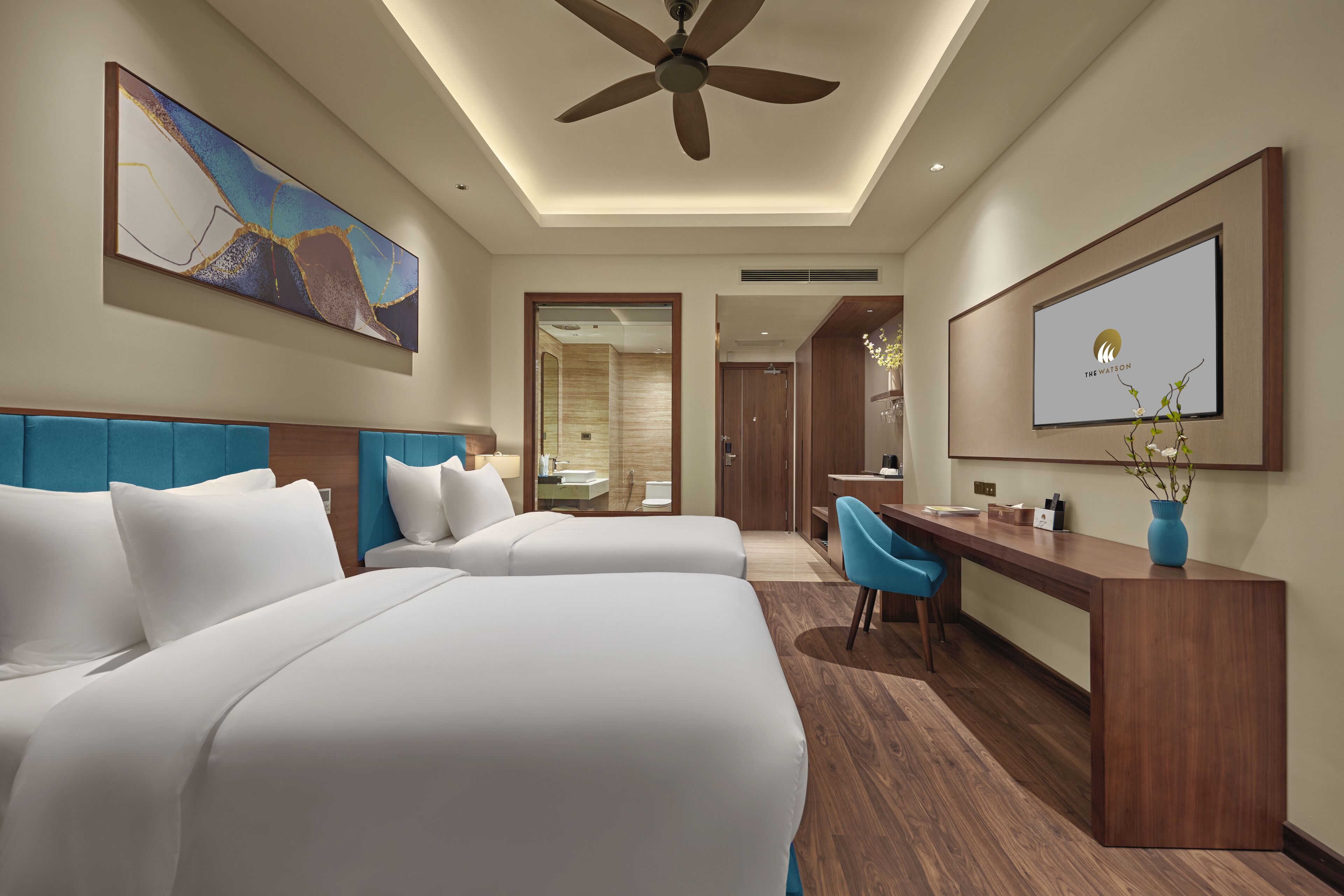 A hotel room with two beds, a desk, a chair, a ceiling fan, and a large abstract painting on the wall.