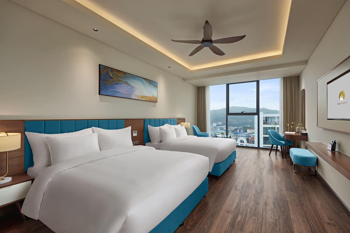 A hotel room with two beds, a desk, a chair, and a large window offering a city view.