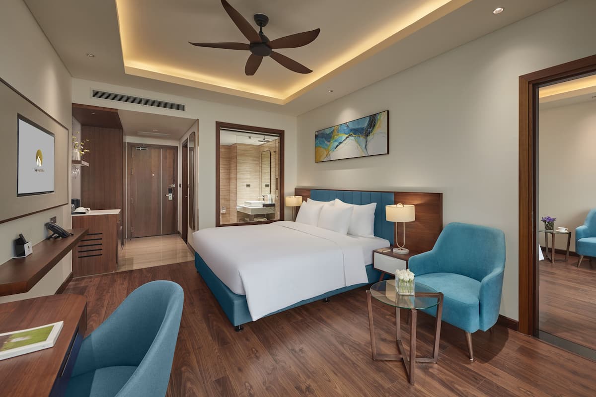 A hotel room with a bed, a desk, a chair, a glass table, a ceiling fan, a painting, and a mirror.