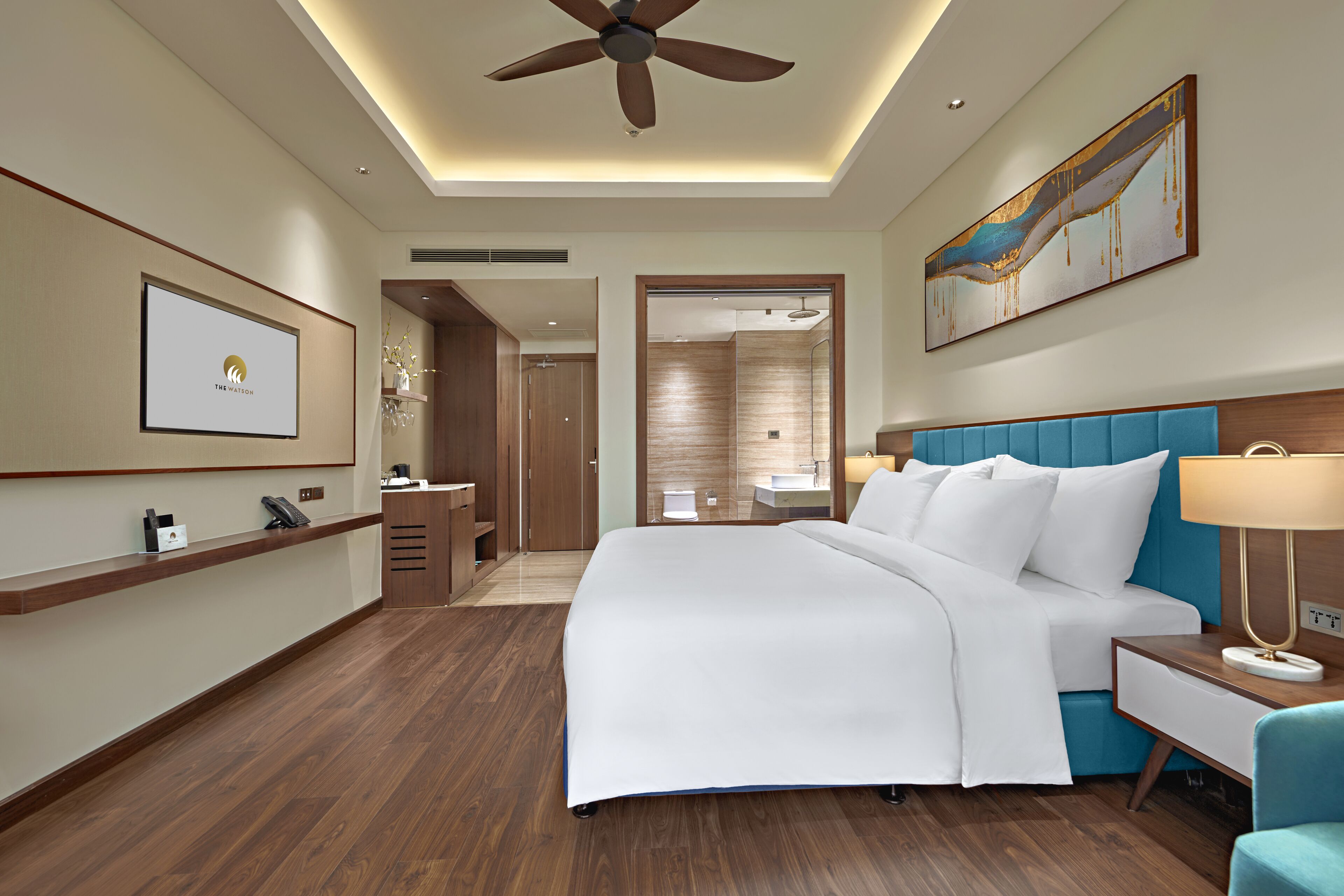A hotel room with a bed, a desk, a chair, a ceiling fan, a painting, and a reception desk.