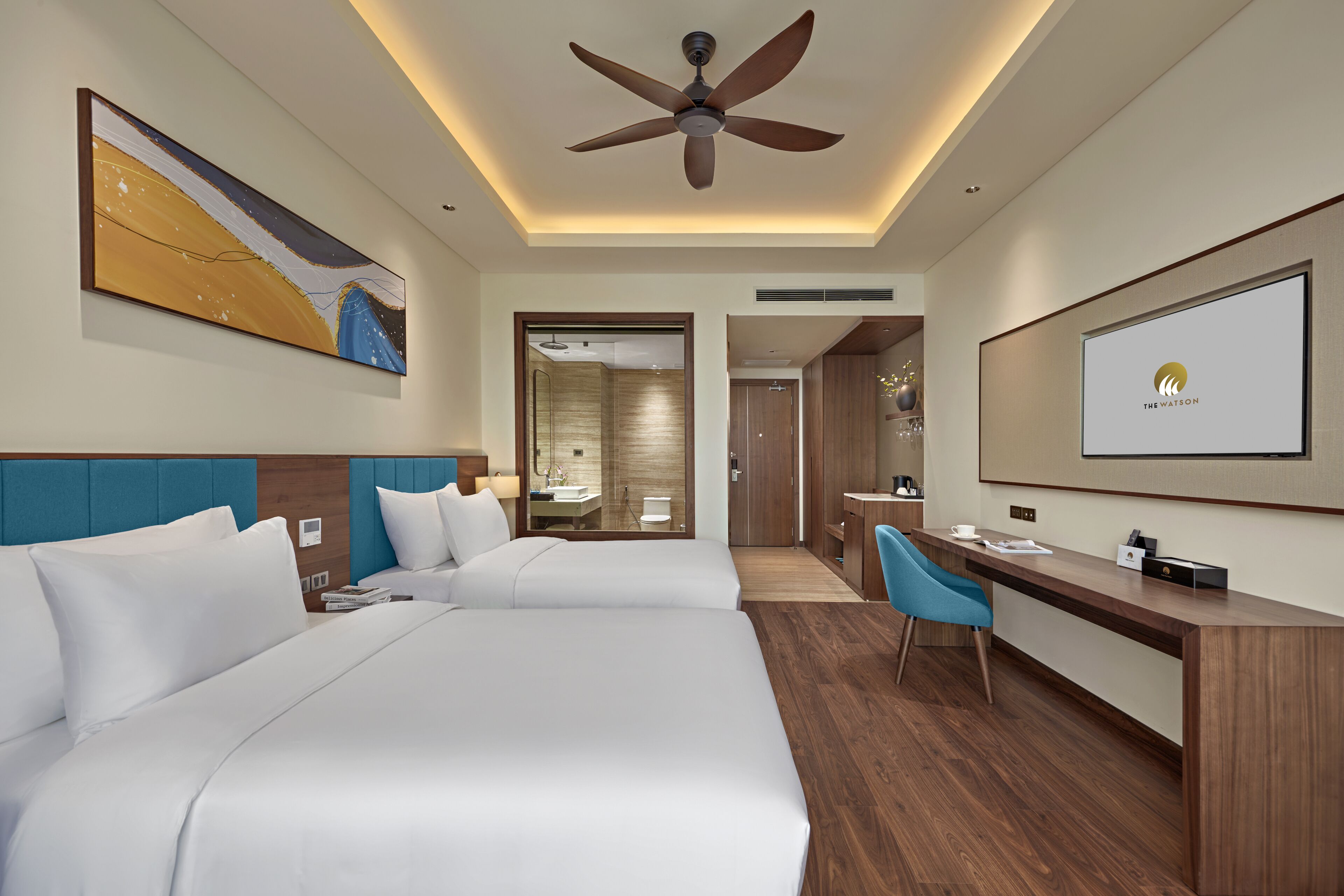 Quarto de hotel com duas camas, uma escrivaninha, uma cadeira, um ventilador de teto e um espelho grande.