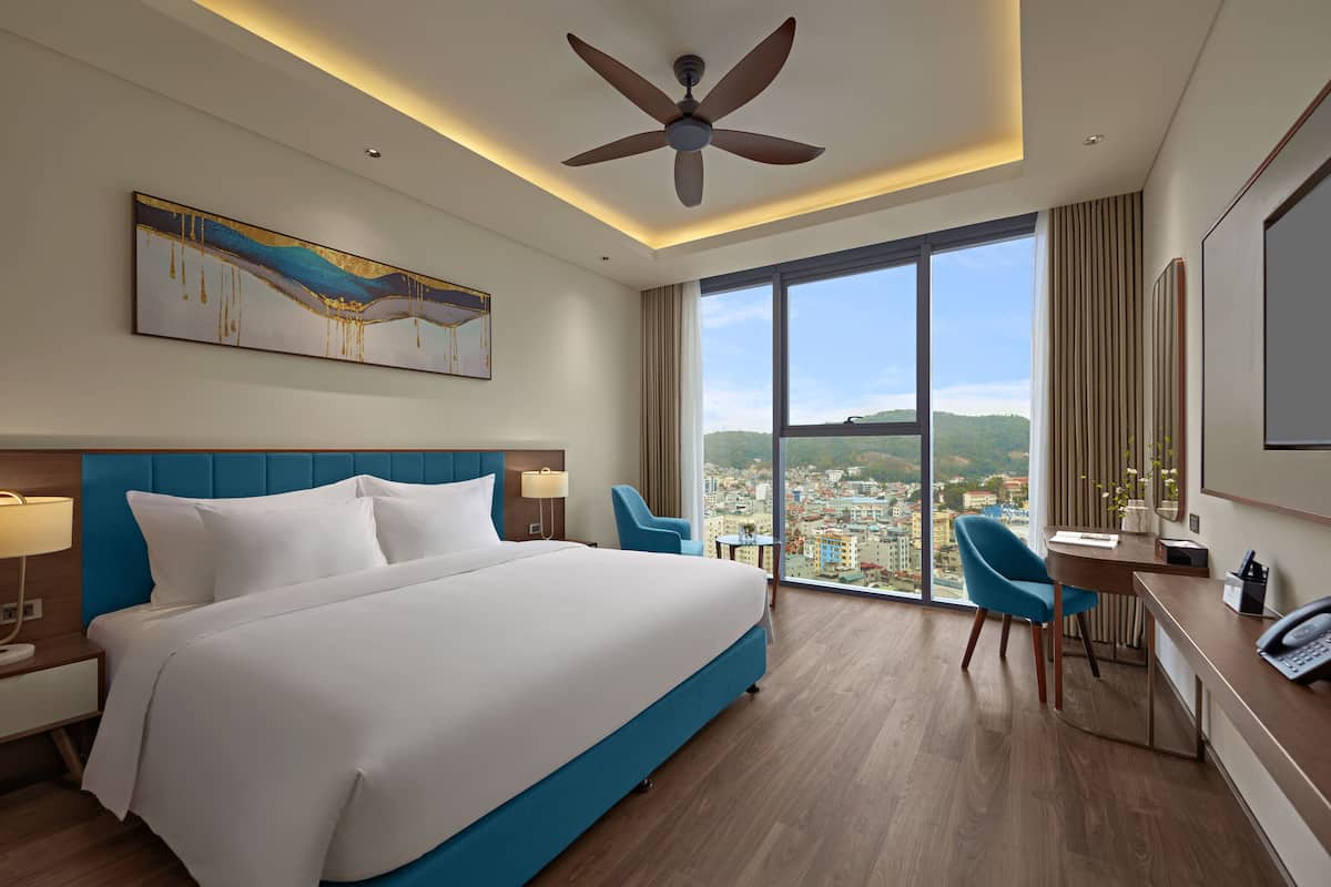 A hotel room with a large bed, a desk with a chair, a ceiling fan, and a large window offering a city view.
