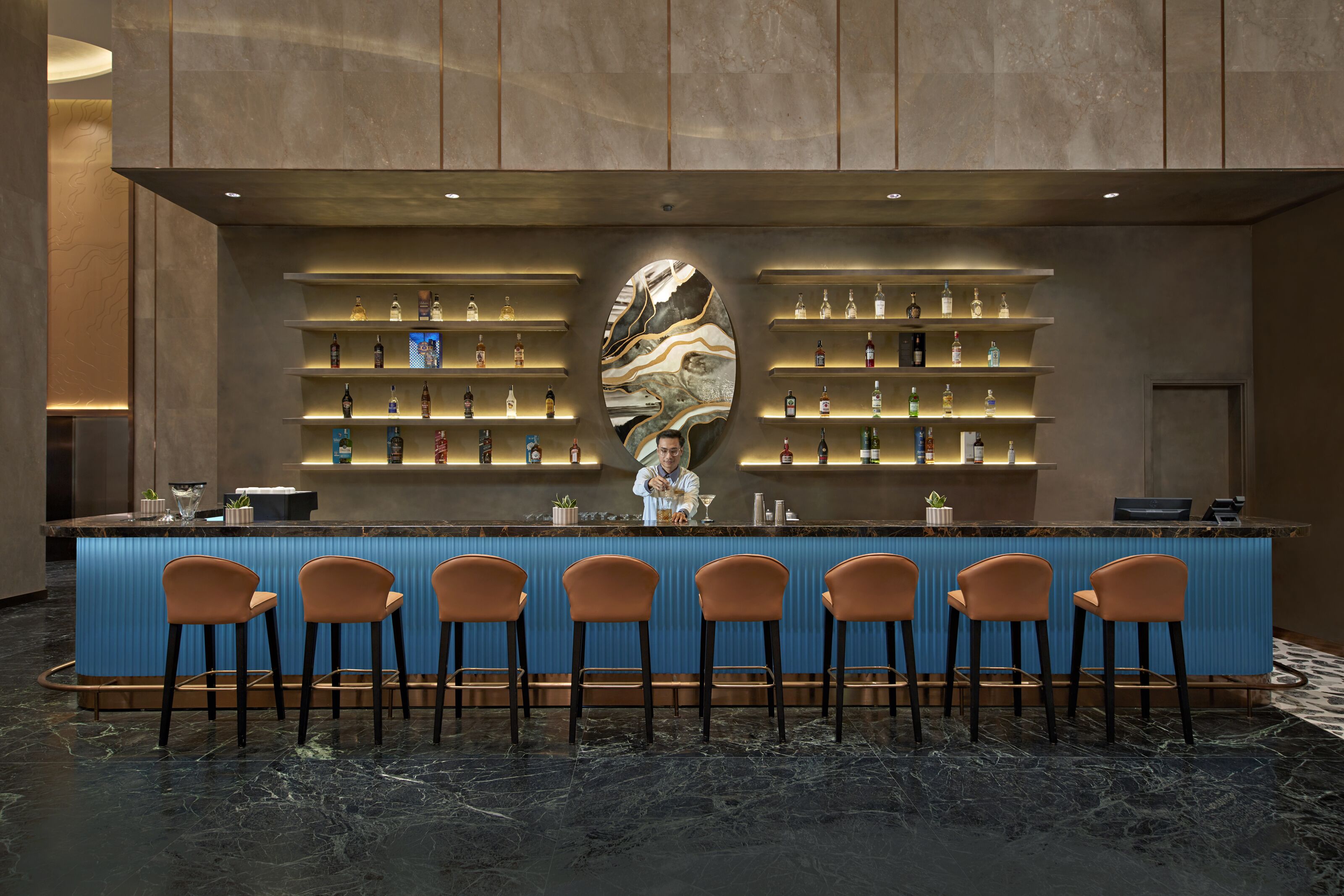 Una zona de bar con una barra azul, varias estanterías llenas de botellas y seis taburetes.
