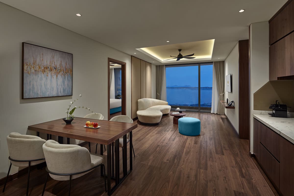 A modern hotel room with a wooden dining table, comfortable seating, a large window with a view, and a ceiling fan.