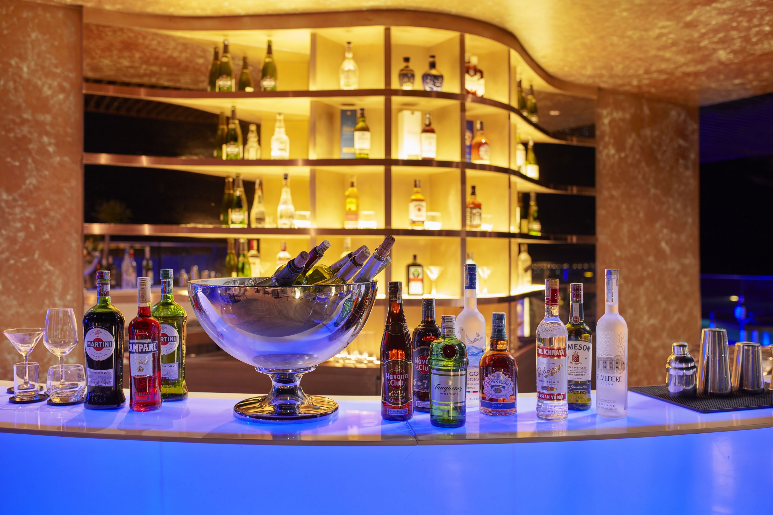 Un espace bar avec une variété de bouteilles d’alcool et de verres, un grand saladier en argent contenant des bouteilles, et une étagère incurvée en arrière-plan.