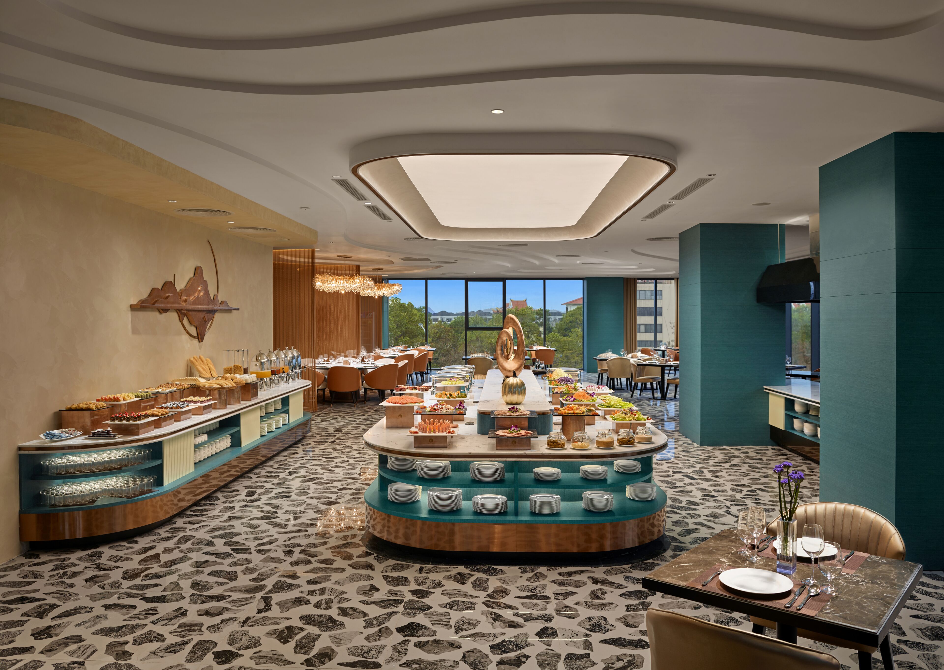 A hotel dining area with multiple buffet stations, a central island with a decorative sculpture, and a view of the outdoors through large windows.