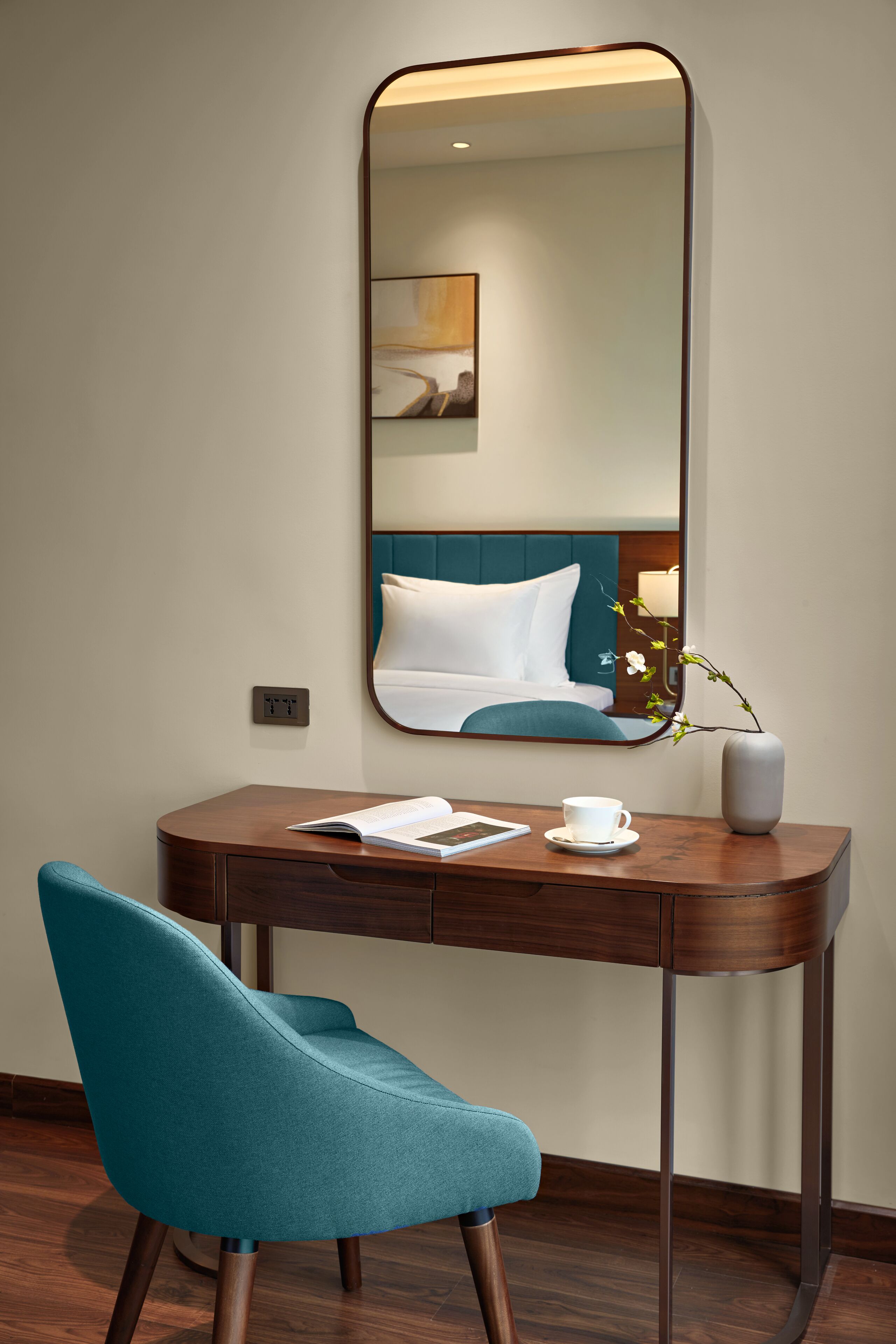 A hotel room with a wooden desk, a teal chair, a mirror, a vase with a flower, and a bed with teal bedding.