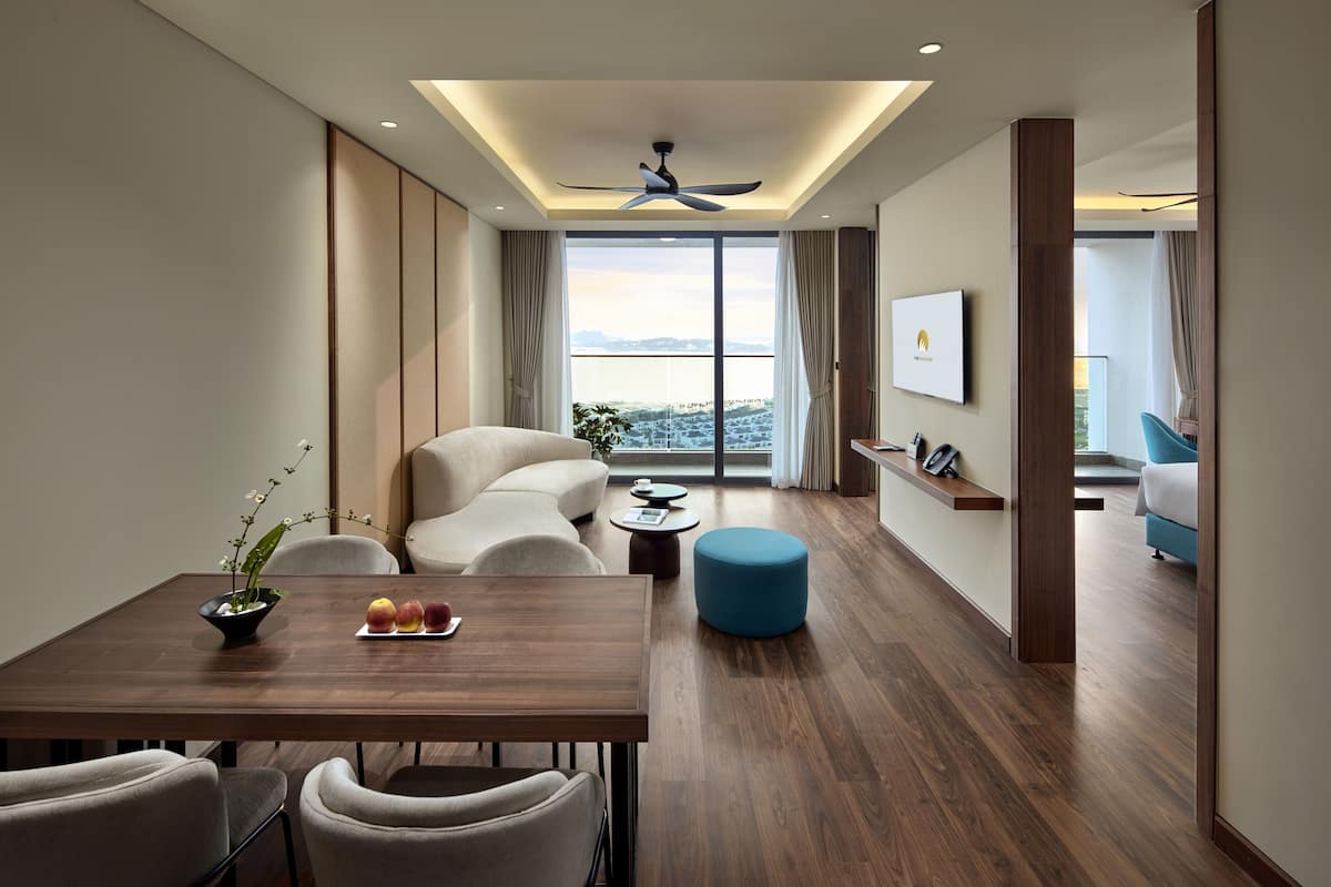 A modern hotel room with a dining area, a sofa, a blue ottoman, a ceiling fan, and a large window with a city view.