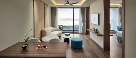 Una habitación de hotel moderna con comedor, sofá, reposapiés azul, ventilador de techo y un ventanal con vistas a la ciudad.