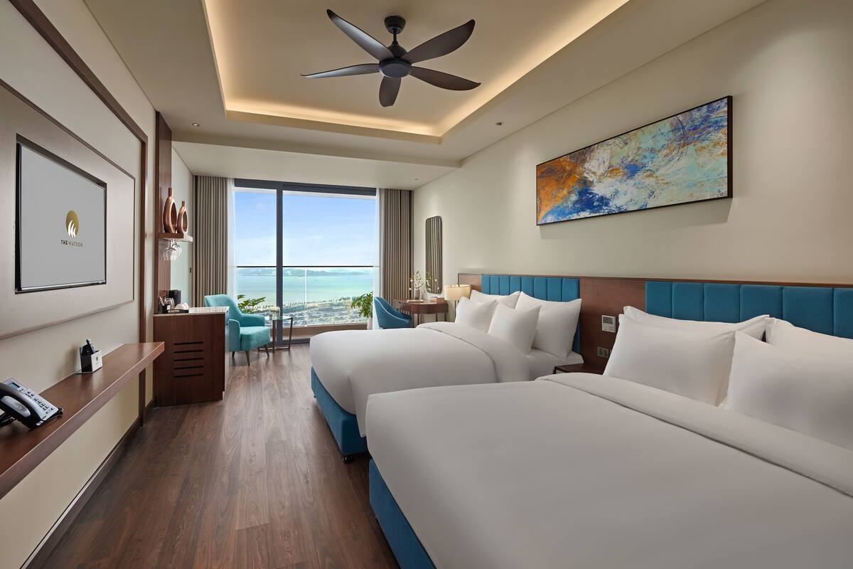 A hotel room with two beds, a desk, a chair, and a large window with a view of the ocean.
