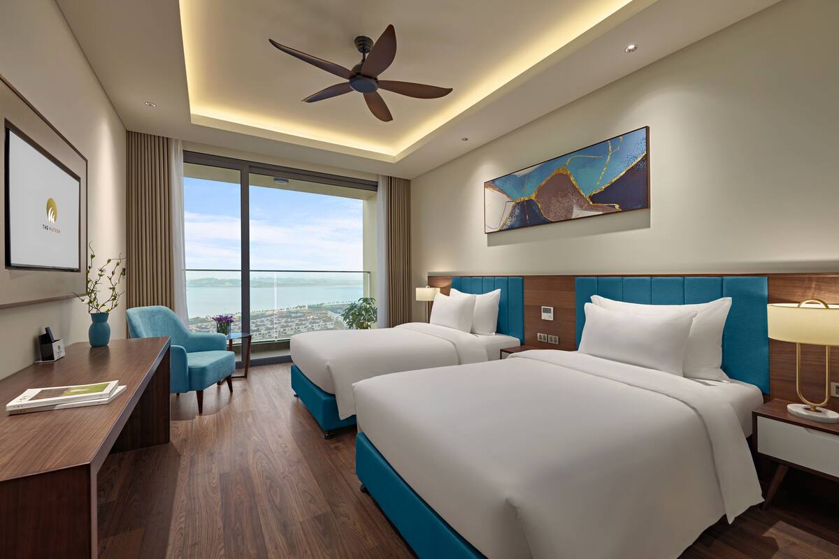 A hotel room with two beds, a desk, a chair, a ceiling fan, a large window with a city view, and a painting on the wall.
