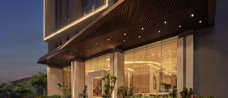 Une entrée d’hôtel moderne avec une façade en verre, un espace extérieur aménagé pour s’asseoir et une façade bien éclairée.