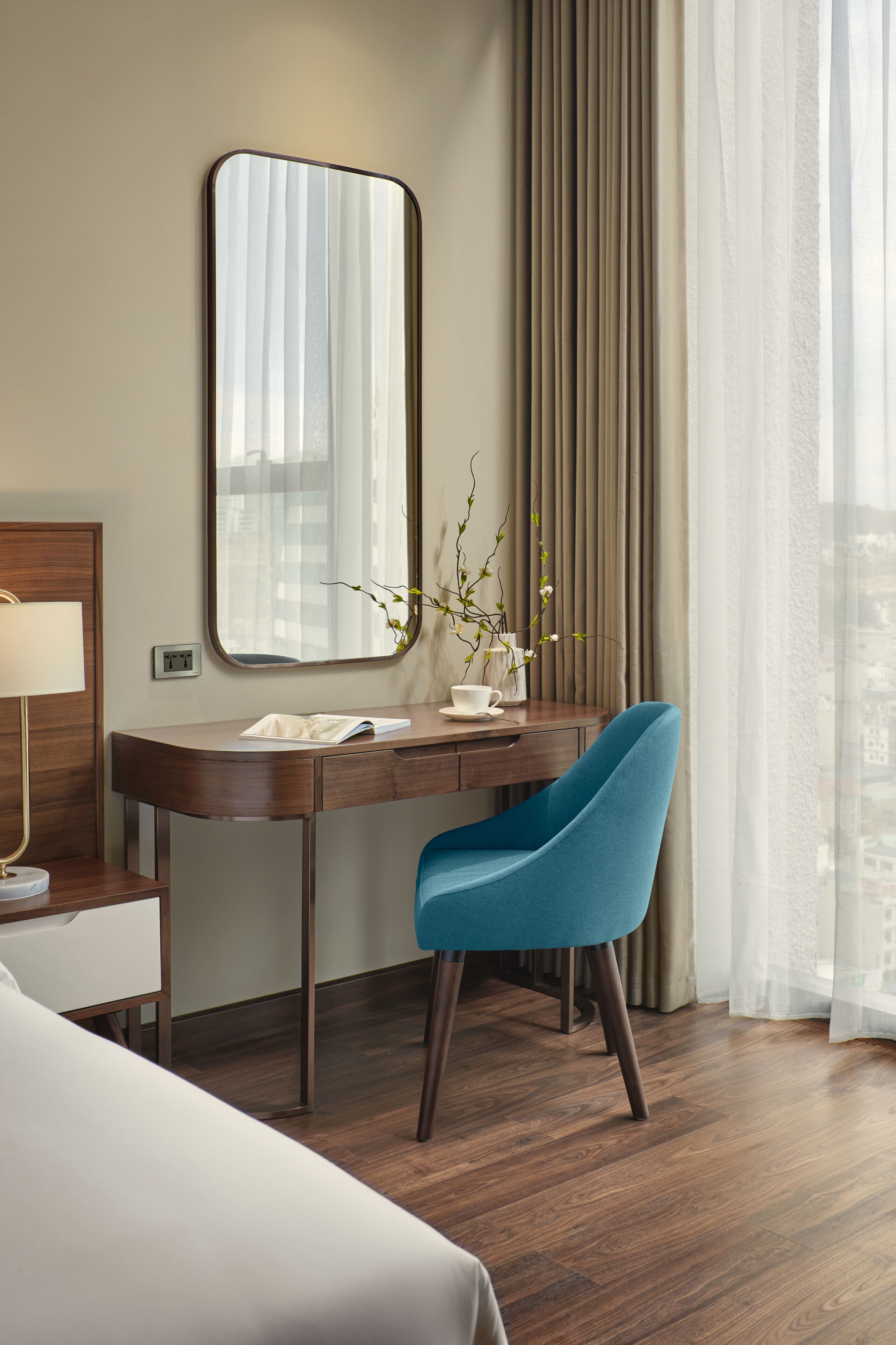A hotel room with a wooden desk, a blue chair, a large mirror, a vase with flowers, and a view of the city through sheer curtains.