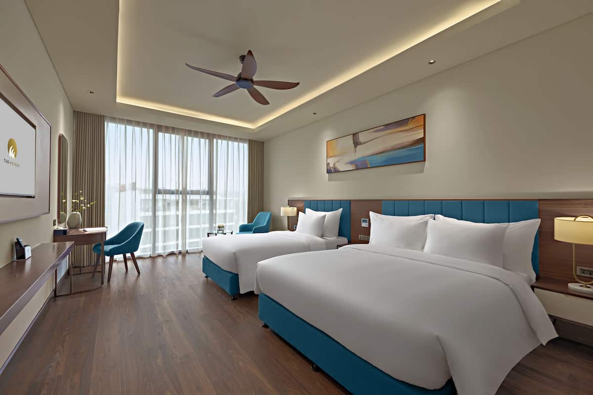 A hotel room with two beds, a desk, a chair, a ceiling fan, and a large window with curtains.