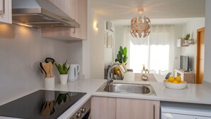 A modern kitchen with a built-in stove, sink, and dishwasher. There is a dining area with a table and chairs, and a living room with a TV and a plant. - Beautiful apartment for 4 guests with pool, A/C, WIFI, TV and balcony (Calafell)