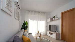 A modern living room with a grey sofa, a coffee table, a TV, and a large window with curtains. - Beautiful apartment for 4 guests with pool, A/C, WIFI, TV and balcony (Calafell)