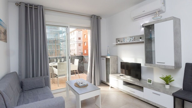 Una sala de estar moderna con un sofá gris, una mesita blanca, un televisor y un balcón con vistas a edificios.