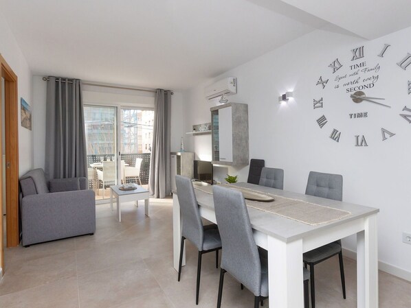 A modern dining area with a white table and grey chairs, a large wall clock, and a balcony with grey curtains.