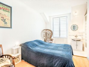 Ein Schlafzimmer mit einem Bett, das mit einer blauen Decke bezogen ist, einem Holzstuhl, einem kleinen Nachttisch, einem Fenster mit Rollläden, einem Spiegel und einem Poster an der Wand.