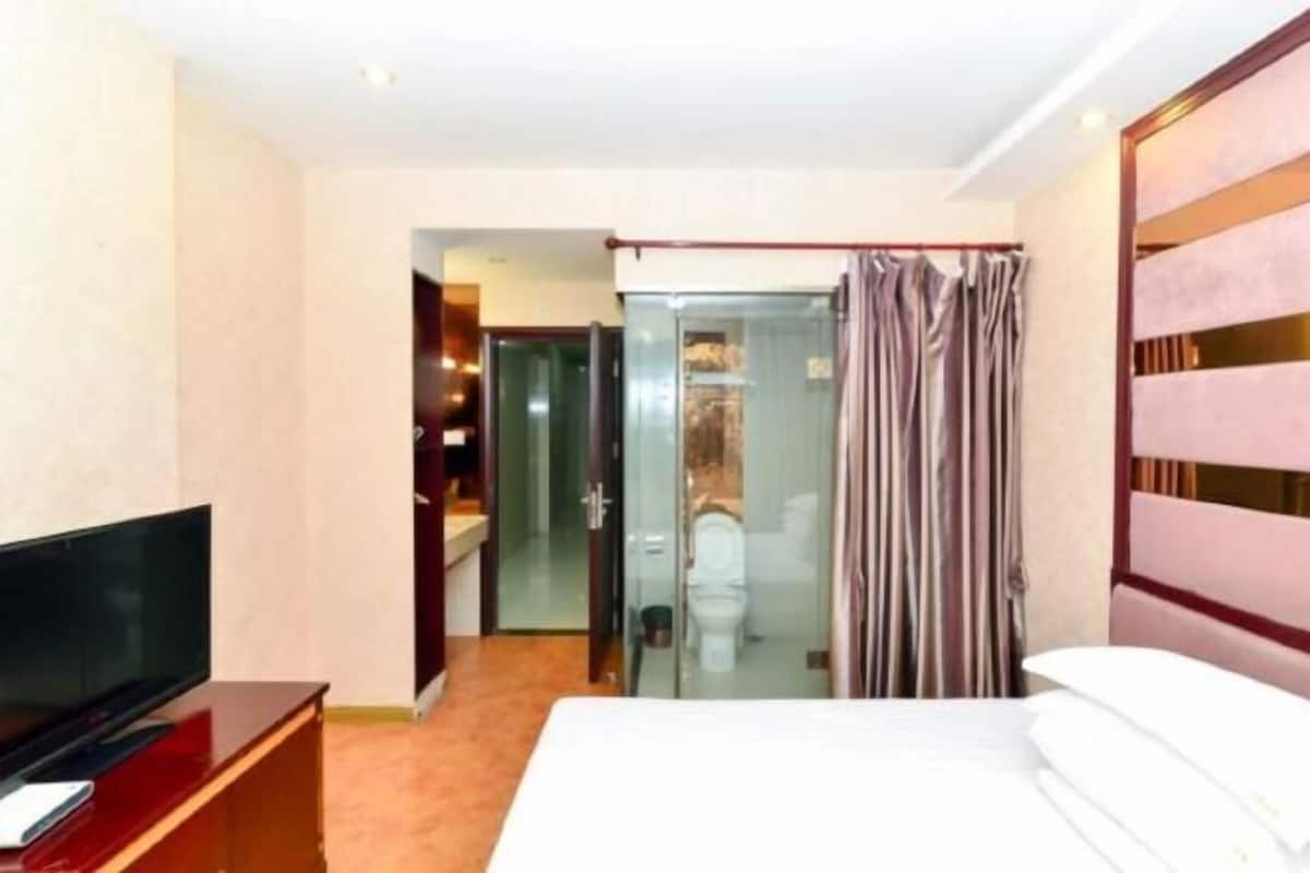 A hotel room with a bed, a TV, a glass-enclosed bathroom, and a wooden wardrobe.