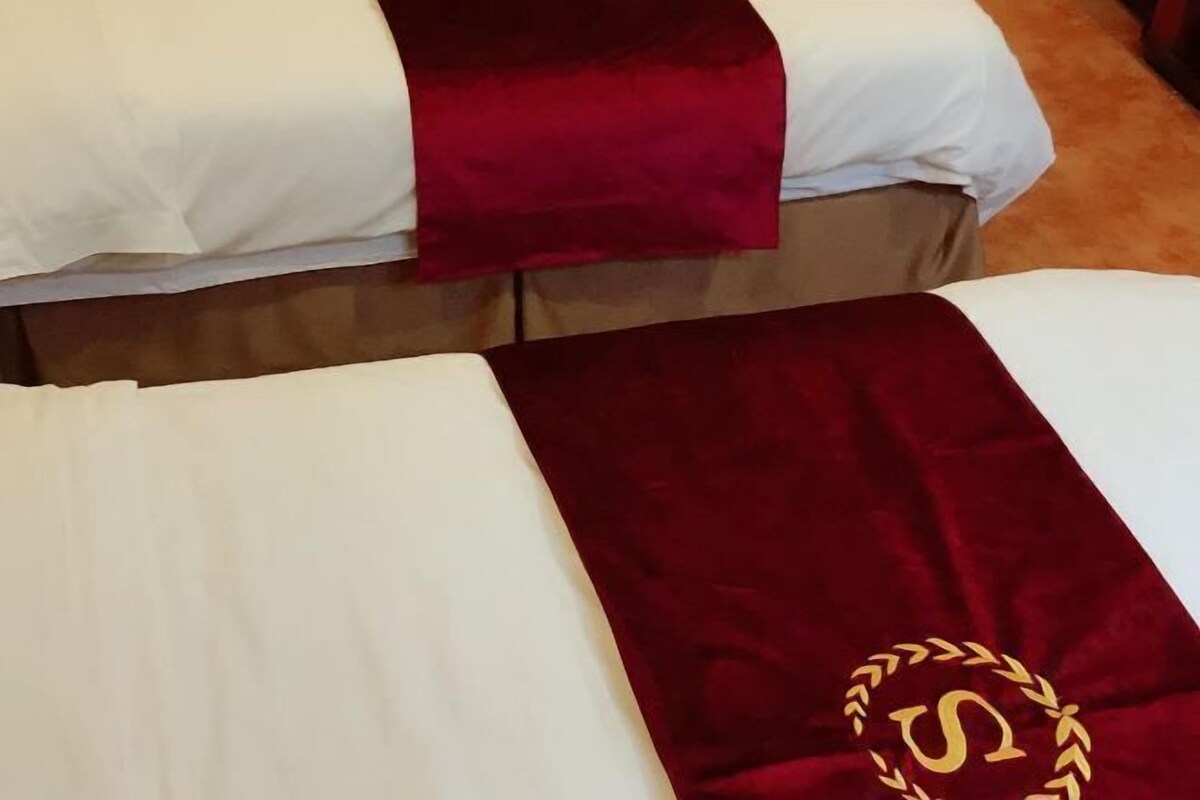 Dos camas con ropa de cama blanca y cabeceras rojas, piso cubierto de alfombra marrón y un emblema dorado en la cabecera.