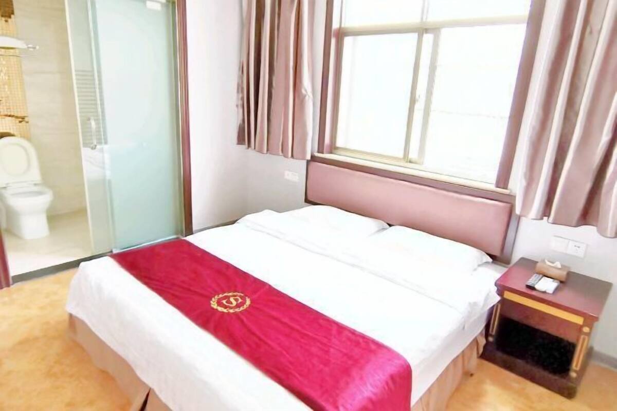 A hotel room with a bed, a bedside table, a nightstand, a window, and a shower area.