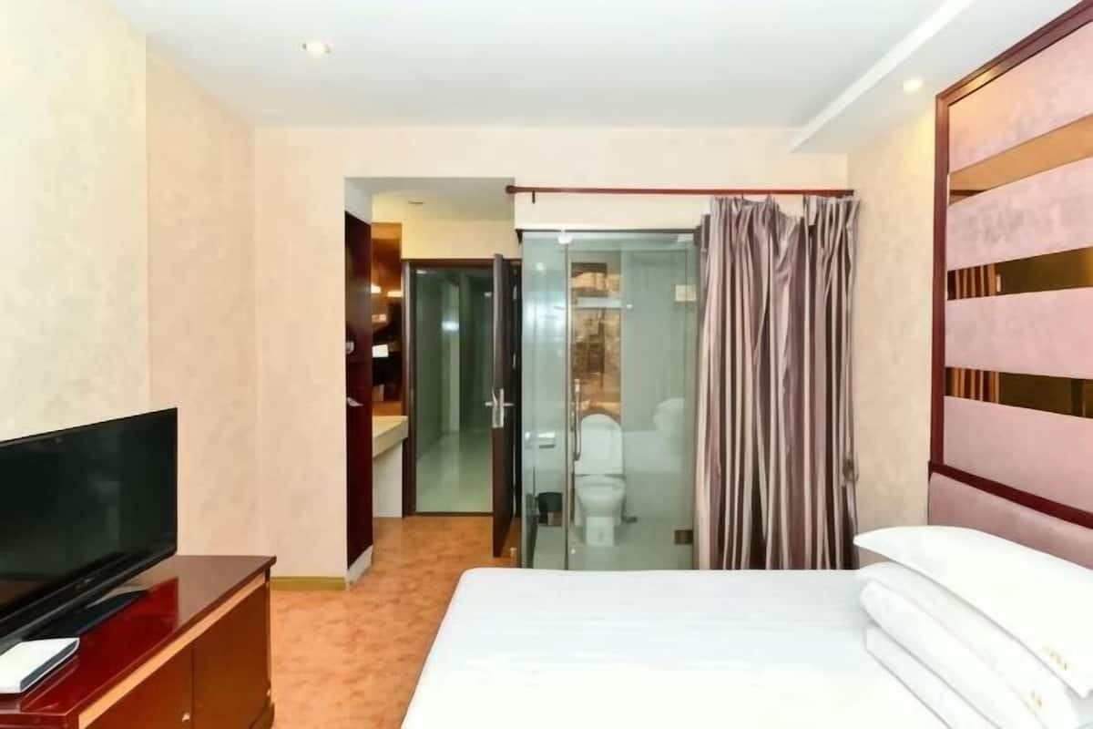 A hotel room with a bed, a TV, a glass shower enclosure, and a sliding door to a bathroom.
