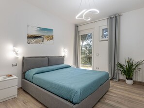 Relax in this serene bedroom with a large bed, a bedside table, and a potted plant, offering a view of the surrounding trees and beach.