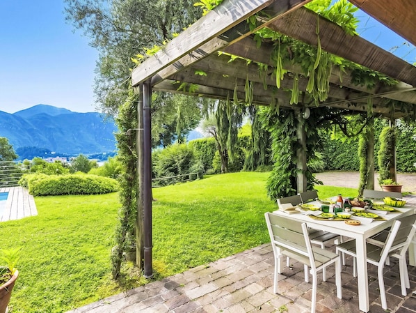 A covered patio with a table set for dining, surrounded by lush greenery and a view of mountains.