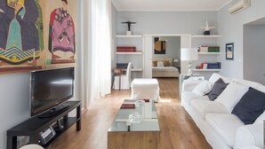 A modern living room with a flat-screen TV, a glass coffee table, a white sofa, and a painting on the wall.