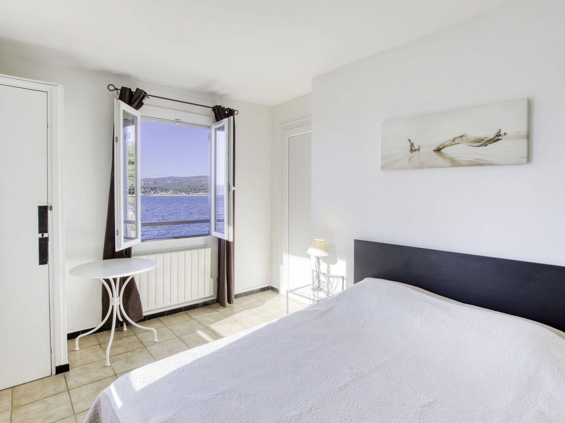 Ein Schlafzimmer mit einem großen Bett, einem Fenster mit Vorhängen, einem kleinen Tisch und einem Bild an der Wand.