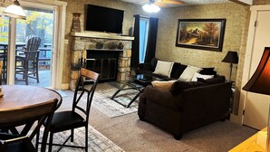 A living room with a stone fireplace, a flat-screen TV mounted above it, a couch, a dining table with chairs, and a ceiling fan. - Relax and unwind Townhome in Snow Ridge Village at Jack Frost mtn in the Poconos (Lake Harmony)