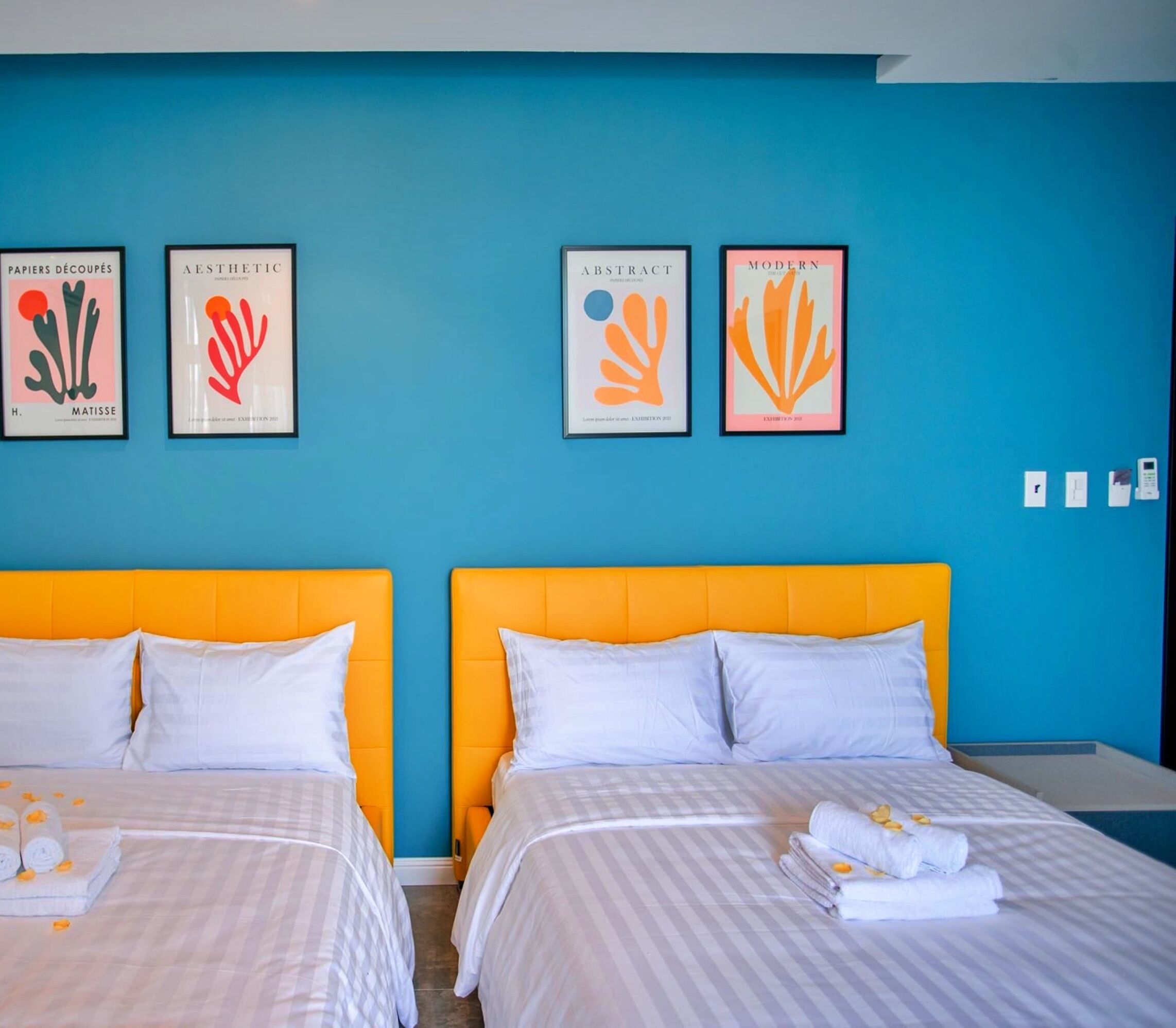 Een hotelkamer met twee bedden, elk met een wit-grijs gestreept dekbedovertrek, en een feloranje hoofdbord. Boven de bedden hangen kunstwerken in lijsten, en op de vloer staat een klein tafeltje met handdoeken en een fles.