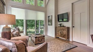 A spacious living room with a large window, a wooden cabinet, a television, and a patterned rug. - Golf and Riverfront Cottage | King Bed | Group Availability (Mineral Bluff)