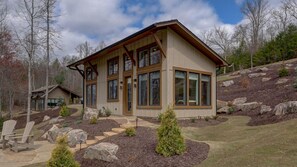 Une maison moderne à deux étages, dotée de grandes fenêtres, d’un chemin en pierre et d’un jardin aménagé avec des rochers et des arbustes.