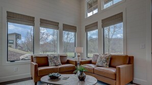 A spacious living room with two leather sofas, a round coffee table, and large windows with blinds. - Golf and Riverfront Cottage | King Bed | Group Availability (Mineral Bluff)