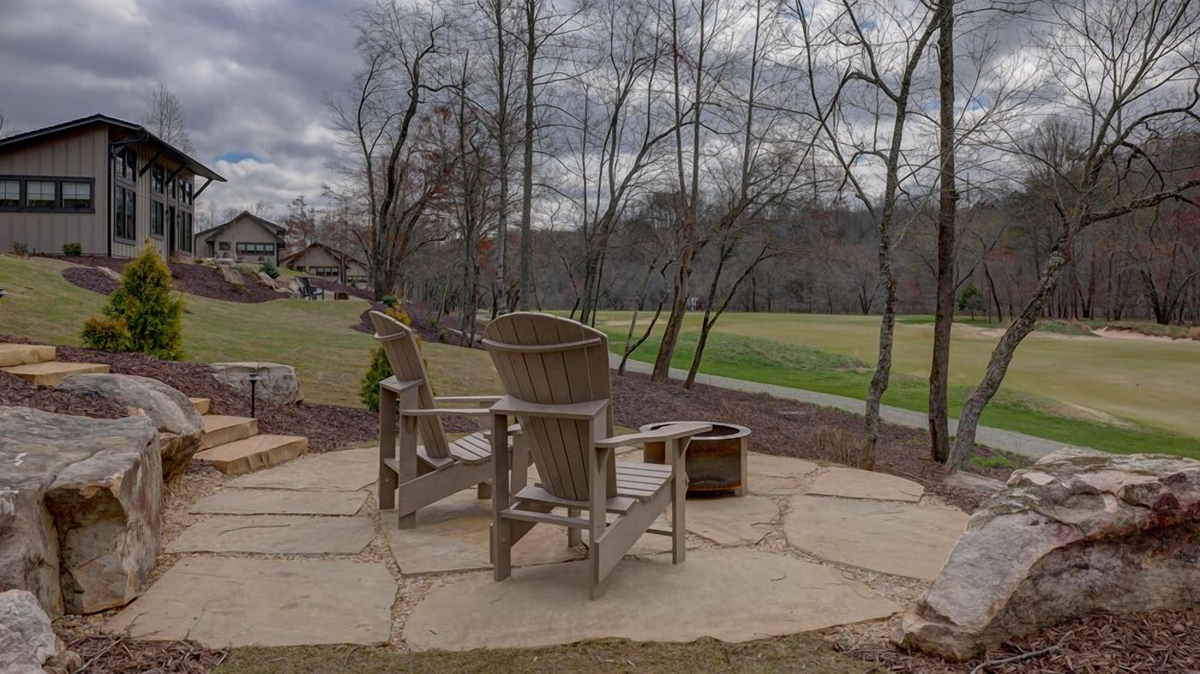 An outdoor seating area with Adirondack chairs and a stone patio, overlooking a golf course and a building with a modern design.
