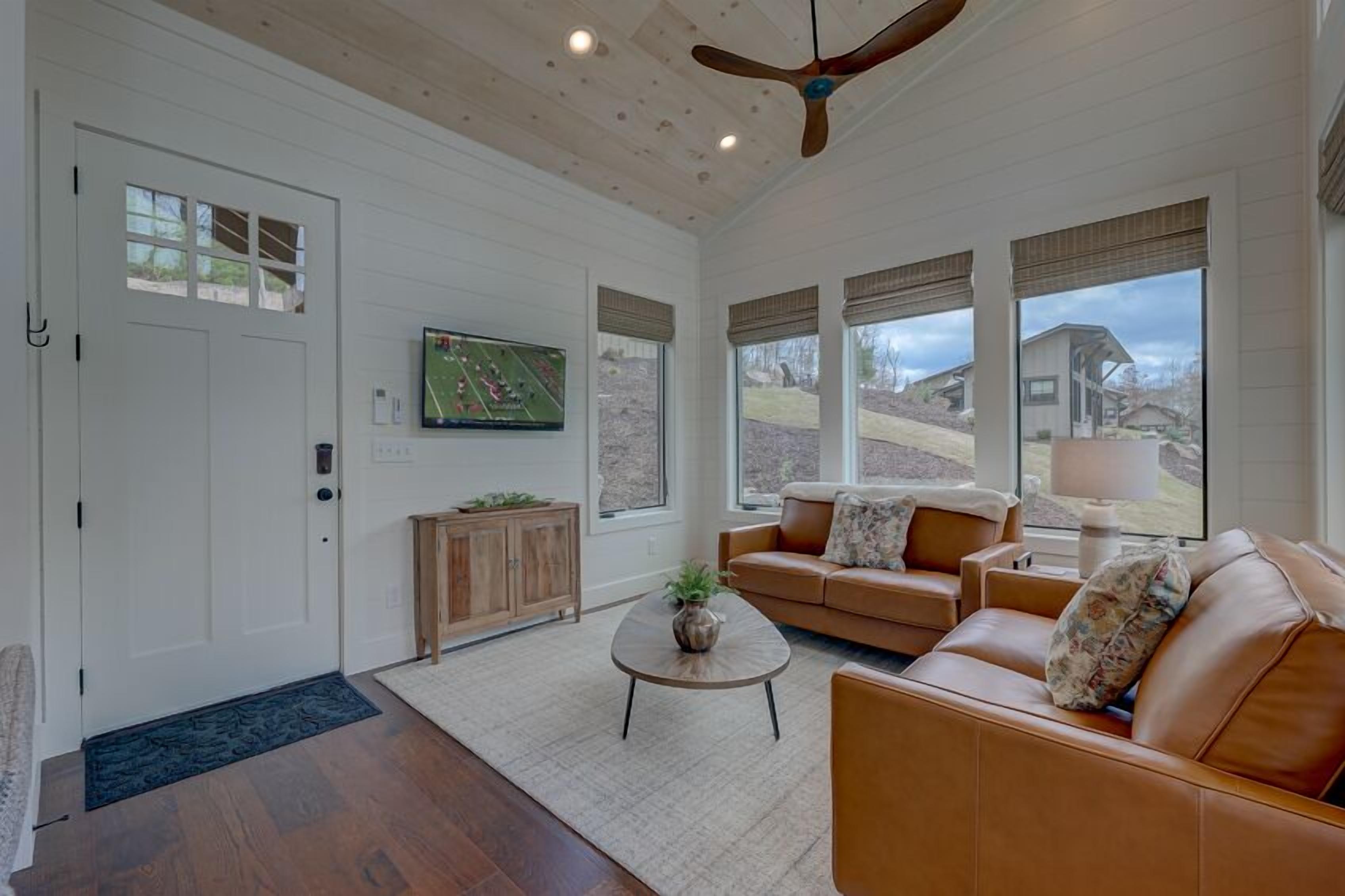 A spacious living room with a large window, a ceiling fan, and a television on the wall.
