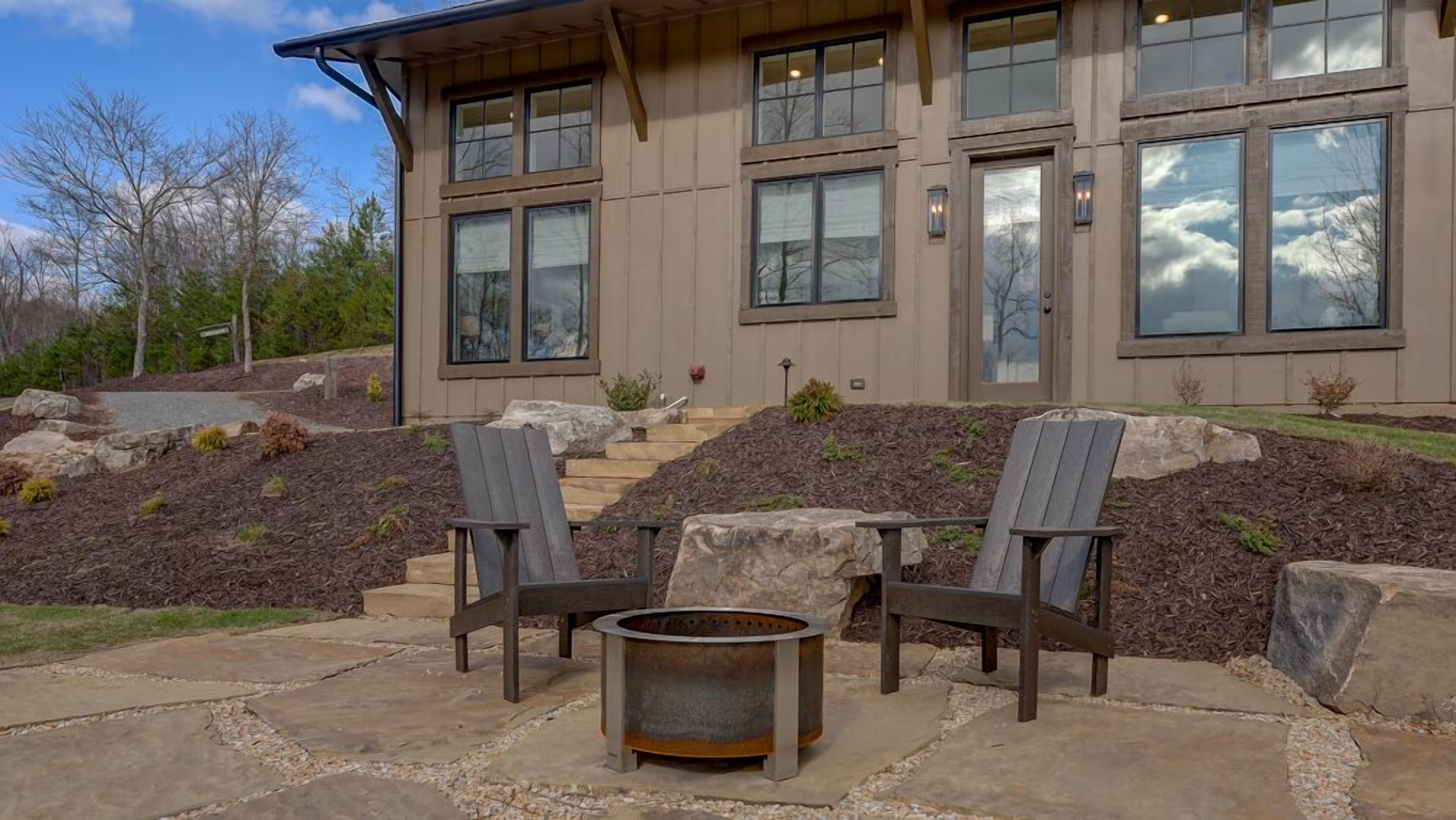 Una casa moderna con un patio de piedra, dos sillas Adirondack y un pozo de fuego.