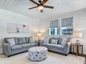 A modern living room with a gray sectional sofa, a round ottoman with nautical prints, a ceiling fan, and a window with patterned valance.