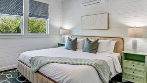 Un dormitorio con cama, dos mesitas de noche, ventilador de techo, aire acondicionado y ventana con vista a follaje.